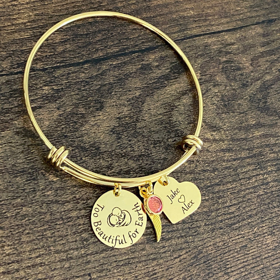 Yellow Gold stainless steel trip loop expandable bangle charm bracelet. first charm is engraved with twin babies under angel wings along with the saying "Too beautiful for earth". next is a heart charm engraved with the names jack and alex next is the twins october birthstone and an angel wing charm.