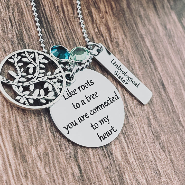 Round pendant necklace engraved with the phrase "like roots to a tree you are connected to my heart". a tree of life charm lays on top of the engraved round pendant. Next to the pendant is a rectangle charm engraved with "unbiological sisters". attached to the main pendant is a may and august birthstone. all charms are attached to a stainless steel silver ball chain.
