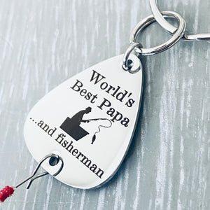 close up view of a 1 inch silver stainless steel fishing lure keychain. Engraved with "World's Best Papa...and fisherman" along with an image of a man fishing from a boat. attached to a durable steel keyring and fishing feather.