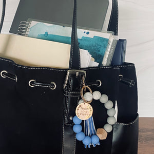 the medium blue, marble, grey, and 1 wood beaded silicone beaded bracelet with the "teach love inspire" rose gold round char tag hooked to a teachers bag