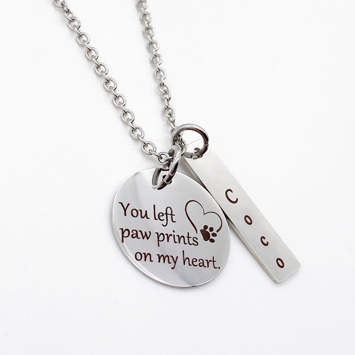 silver 3/4 inch pendant engraved with "you left paw prints on my heart" with an image of a heart and paw print. Next to it is a rectangle tag personalized with the name Coco. Pendants are attached to a cable chain.