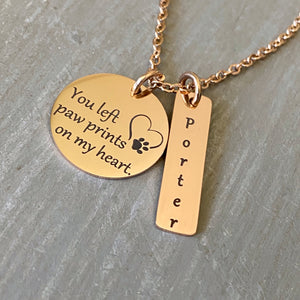 Rose gold 3/4 inch pendant engraved with "you left paw prints on my heart" with an image of a heart and paw print. Next to it is a rectangle tag personalized with the name Porter. Pendants are attached to a cable chain.