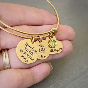 Woman holding a yellow gold triple loop wire bracelet with a round 3/4 inch engraved with "your first breath took mine away", a 5/8 inch round disc with engraved name "Ian" with an image of a mom holding a baby, and a 3/4 inch heart engraved with the birthday date "8.04.17", and an august birthstone.