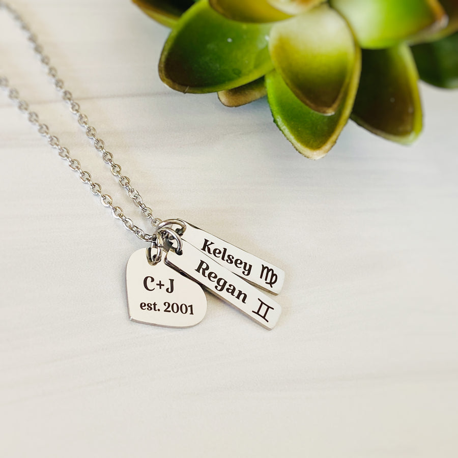Silver mom necklace with heart charm engraved with mom and dad initials "C+J est 2001" with 2 name tags. Regan with a gemini sign. Kelsey with a virgo zodiac symbol