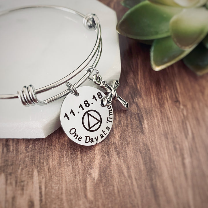 silver triple loop bangle charm bracelet with a 7/8" round charm disc engraved with the quote, "One Day at a Time" a AA symbol and personalized sobriety date 11.18.18. Next to the main disc is a silver religious cross